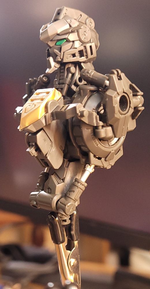 Barbatos in the midst of construction.  The torso and head have been assembled,  held aloft on a rubberized crocodile clip. The mech is a mass of grey plastic,  accented by a golden crest on its chest and the signature emerald eyes of a Gundam.