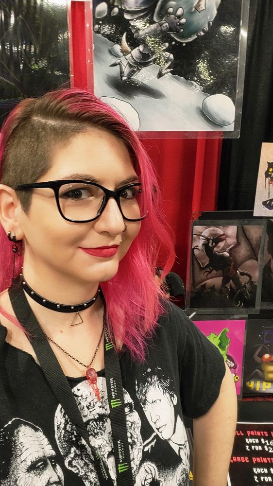 Tiny Neenja, a pink haired side shaved nb person with a black tshirt, smiling a little concerned about the heat in the vendor hall