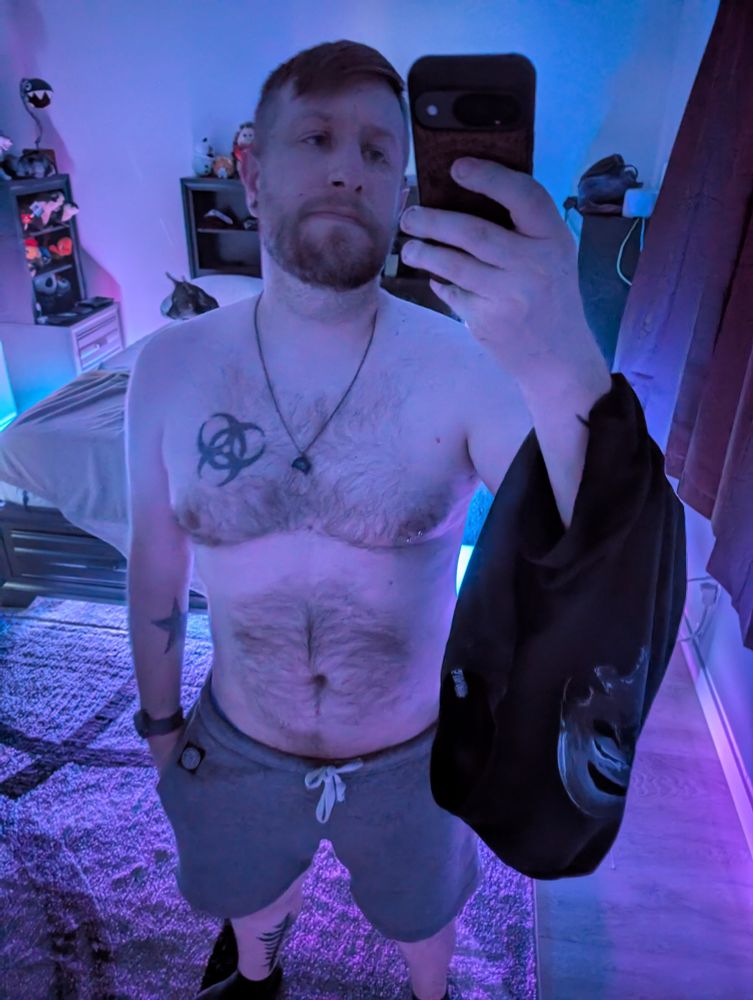 Standing in my bedroom with only the RGB under lighting. I have grey shorts on and a black shirt slung over my arm. I look so, SO tired.