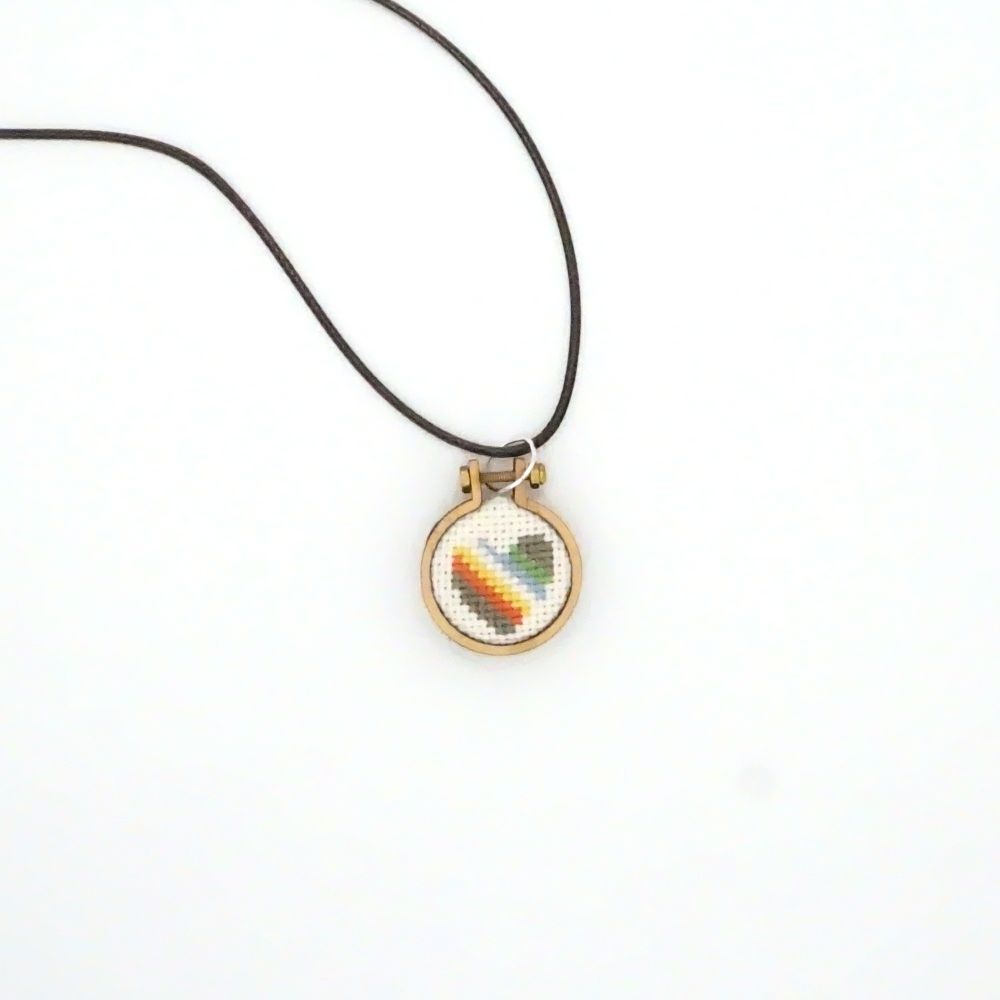 A photograph of a 1" pendant laying on a white background. It's the Disability pride flag cross-stitched and encased in a very small hoop. The flag colours are a muted dary gray, red, yellow, white, light blue and green.