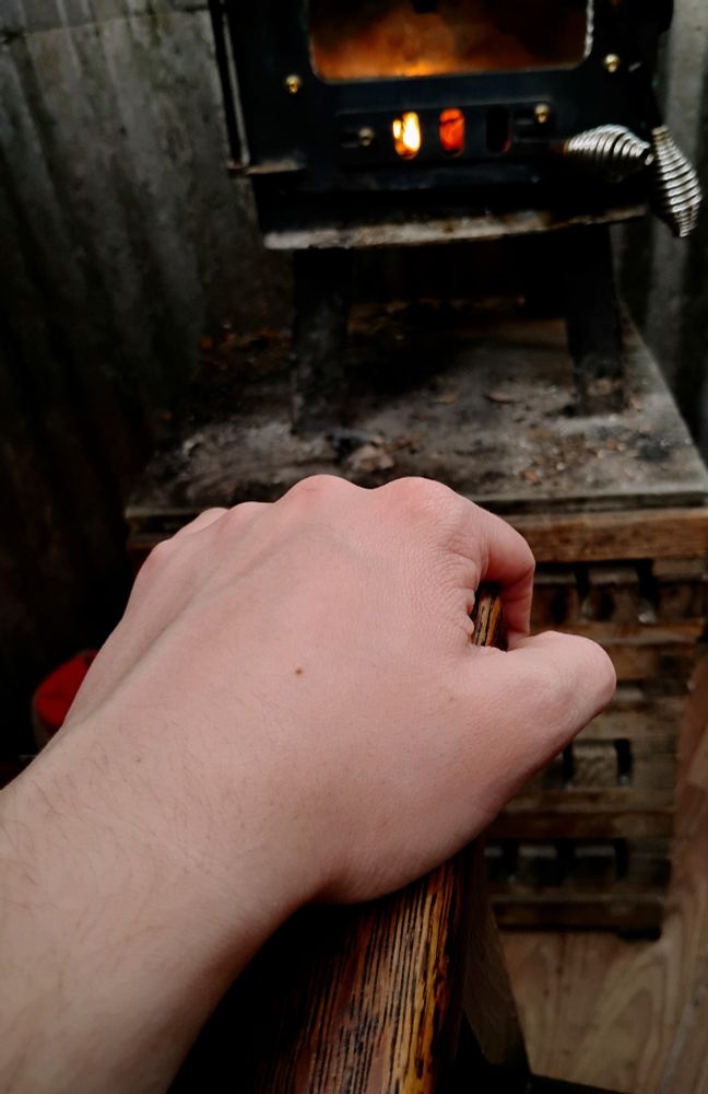 Photo of a hand gripping the wooden armrest of a chair, veins visible on the back of the hand, and a wood stove fireplace in the background. 