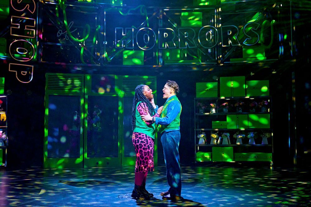 Georgina Onuorah (Audrey) and Colin Ryan (Seymour) in Little Shop of Horrors. Photo Manuel Harlan. Two people stand close together and hold each other as they sing. They wear colourful clothing and are surrounded by specks of green light, cast from an unseen disco ball. Green boxes and cabinets fill the background. 