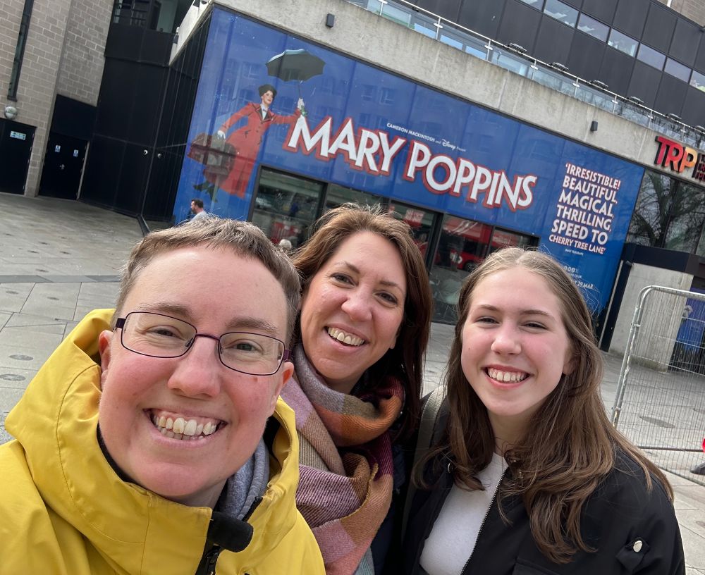 Selfie of Emily, Tabitha and teenage girl outside a theatre. There is a large banner above them that reads “Mary Poppins”.