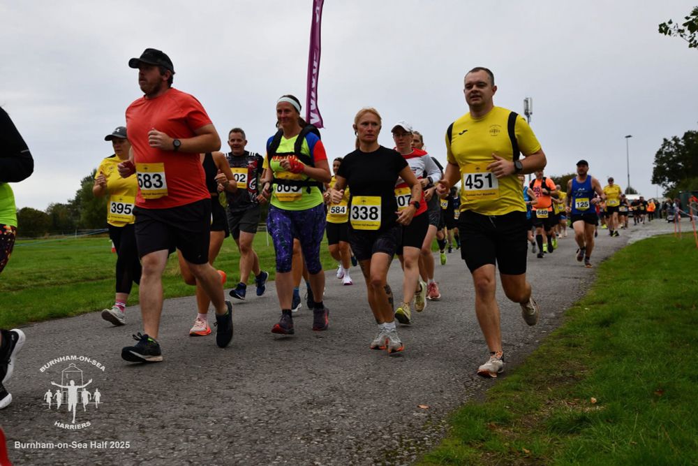 Photo of a group of runners. There is a 2:15 pacer (running with a big flag). Emily is just behind them in her red and white club running top and white cap.