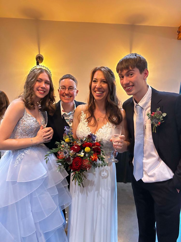 A family photo. The bridesmaid has a huge Cinderella style dress, I am in a three piece black suit and our teenage boy is also in a suit. Tabitha is in a stunning white dress, lace topped and sleeveless. She is holding a bouquet of rainbow inspired flowers.