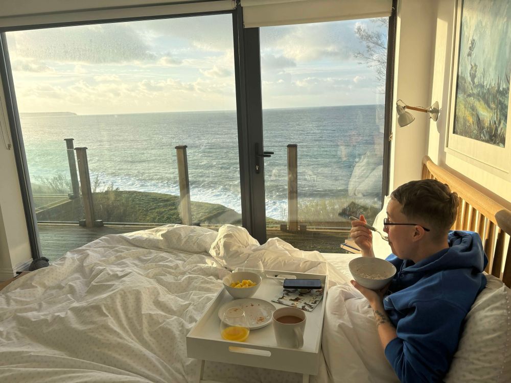 Photo of Emily eating breakfast in bed looking out over the sea.