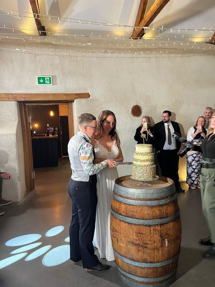 Emily and Tabitha are cutting a three tiered wedding cake, covered in sprinkles, that is sat on top of a barrel.