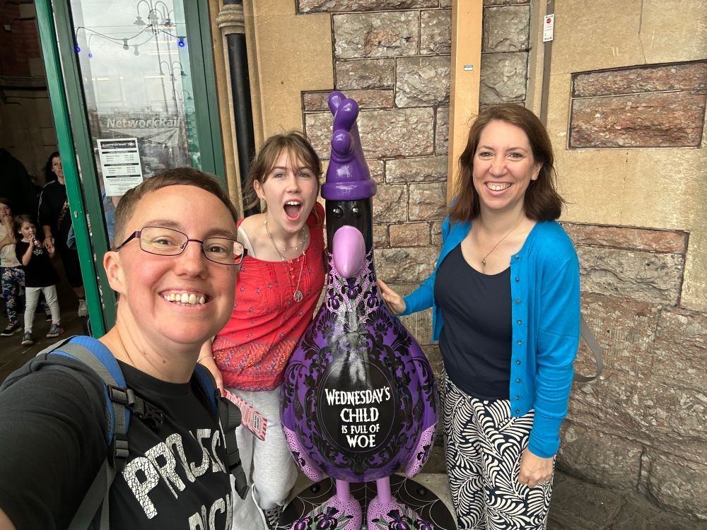 Selfie of Emily and Tabitha with a teenage girl standing next to a model of a penguin wearing a rubber glove on his head and painted themed “Wednesdays Child is full of woe”.