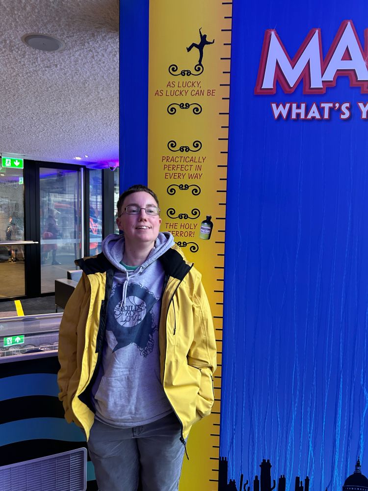 Emily is standing next to a large image of Mary Poppins measuring tape. She measures between “the holy terror” and “practically perfect in every way”.