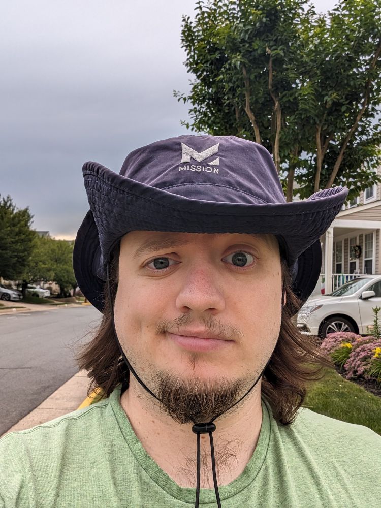 A photo of me with long hair, wearing a green shirt and a dusky blue hat, cocking an eybrow with a quizical, doubtful look