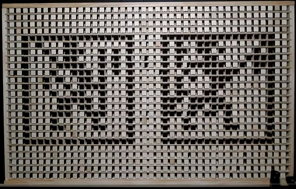 A 40×25 grid of wooden blocks held between horizonal slats. They rotate horizontally: some are rotated to show a black face, others a white one. It is lit with a direct light. Two 16×16 pixel-art images appear side-by-side on the grid: they both have a black border and show two 13-pixel-high human figures facing right: the Infantry Huscarl (on the left) and the Thane (on the right) from Challenge Software's 1989 game /Vikings/ https://worldofspectrum.org/archive/software/games/vikings-challenge-software . They are both stylised images which seem to show men with large round shields and long pole weapons walking right.

The Infantry Huscarl on the left appears to hold a two-handed axe vertically, at about the middle of the shaft, while some kind of spear or javelin appears to be leaned back resting on his shoulder. (A couple of pixels at the middle of the axe head are off when they were on in the original pixel image.) There seems to be a diagonal stripe running top-left-to-bottom-right running through the middle of his shield.

The Thane seems to be holding a two-handed axe forward at a 45° angle while maybe holding some other kind of halberd or other pole weapon vertically behind him. There seems to be a dot, or maybe a boss, off-centre on the shield.

The arm which manipulated the blocks is just visible at the bottom right of the image.