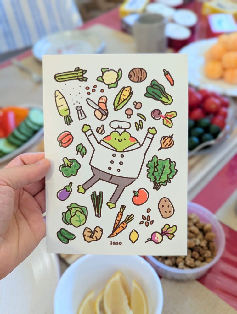 photo of a print of my illustration — of a happy Tom the Frog in their chef outfit with Olivia holding a dull knife and vegetables scattered around