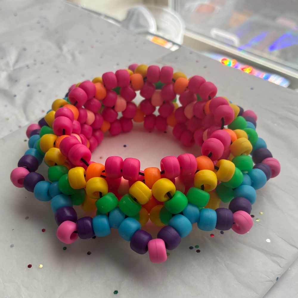 A side angle view of a large kandi bracelet made from matte multicolored pony beads. In the center of the bracelet that wraps around your wrist it is a bright magenta with salmon accents on the interior. From the magenta outwards the colors move in a rainbow pattern including orange, yellow, green, blue, purple, and pink. The bracelet tapers down into a mandala pattern forming soft spike shapes at the points. 