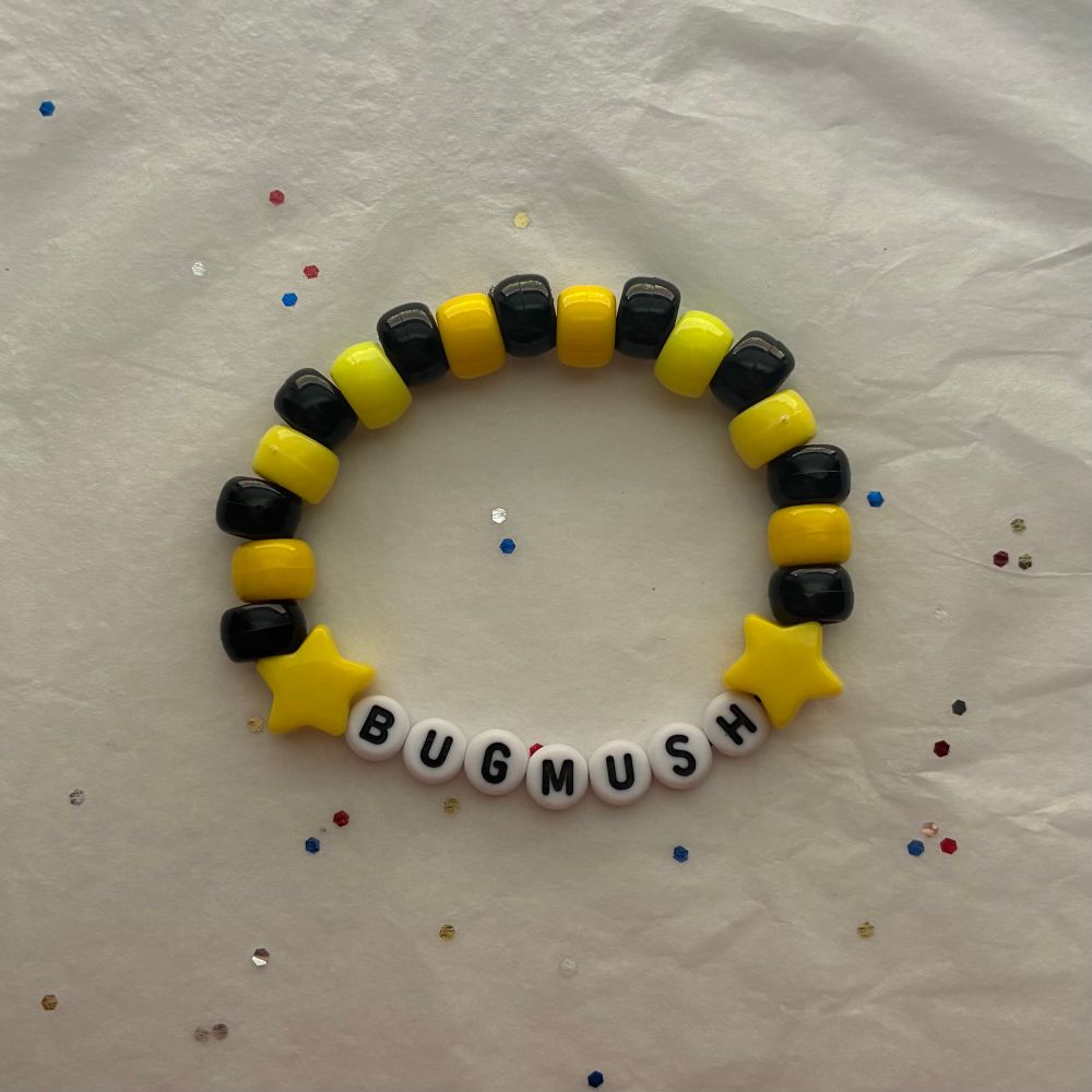 A beaded single kandi bracelet with black and yellow standard round beads as well as two yellow stars surrounding letter beads that spell out the word bug mush. The yellow beads surrounding go in a varied gradient of yellows. 