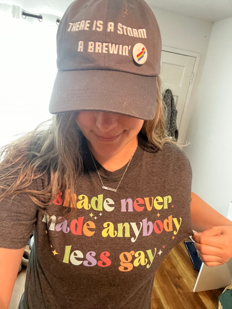 Woman wearing a hat that says “there is a storm a brewin” and a shirt that says “shade never made anybody less gay”