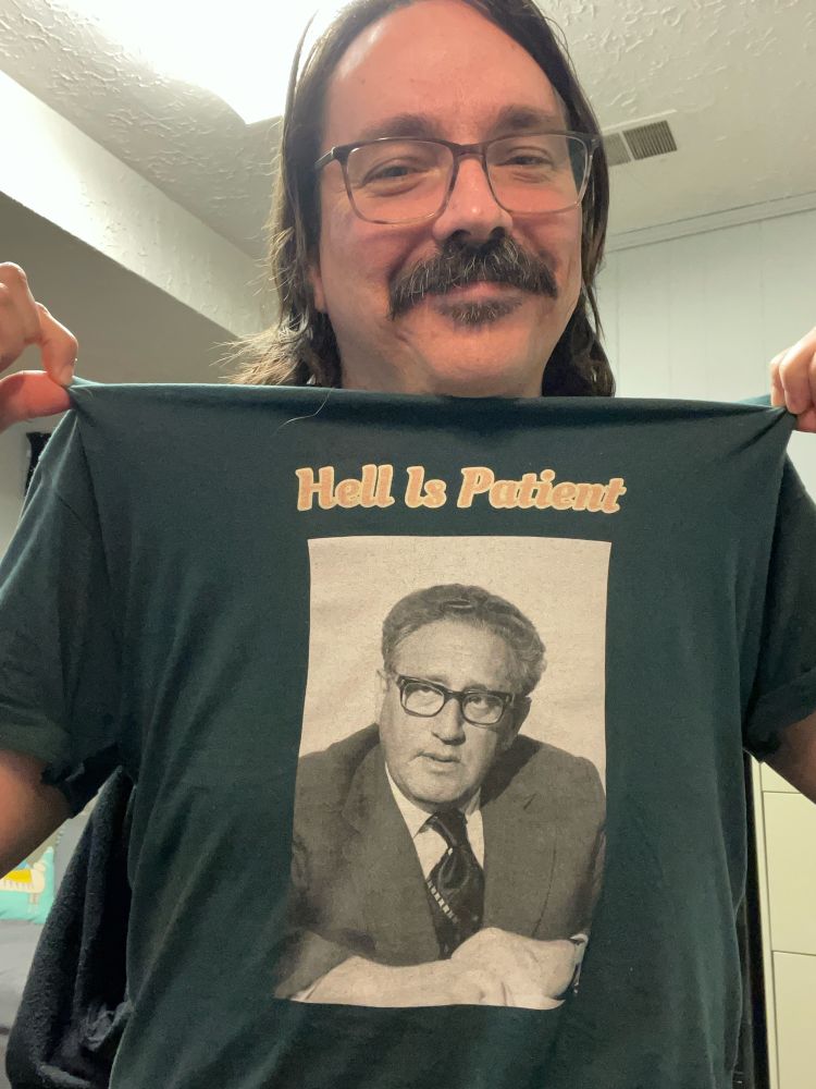 A photo of Billy Wraith Cyrus wearing and showing off a green t-shirt. On the t-shirt are yellow bubble letters that read "Hell is patient" and below the words is a black and white photo of Henry Kissinger.