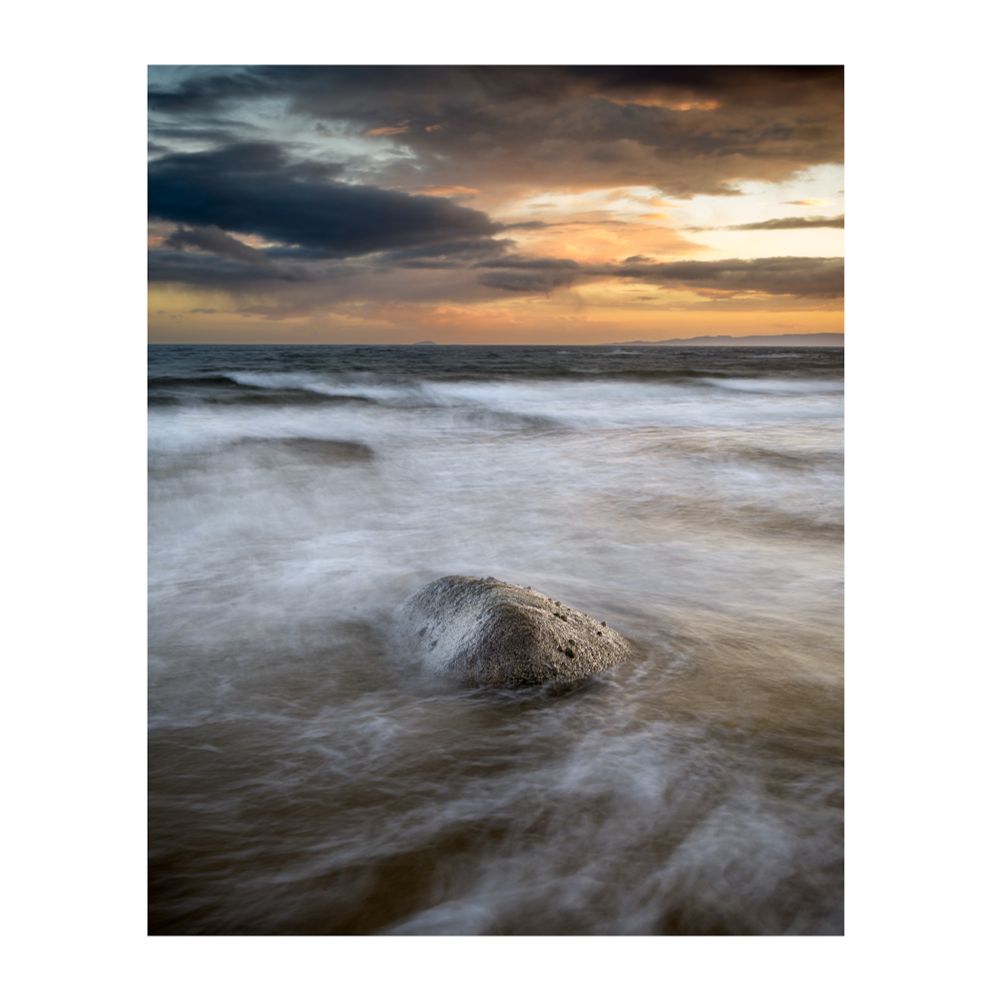 Vertical image of a central small rock surrounded by the swirling motion of the sea, with white foam highlighted by the soft yellow and orange glow coming from a painterly sky and the setting sun. 