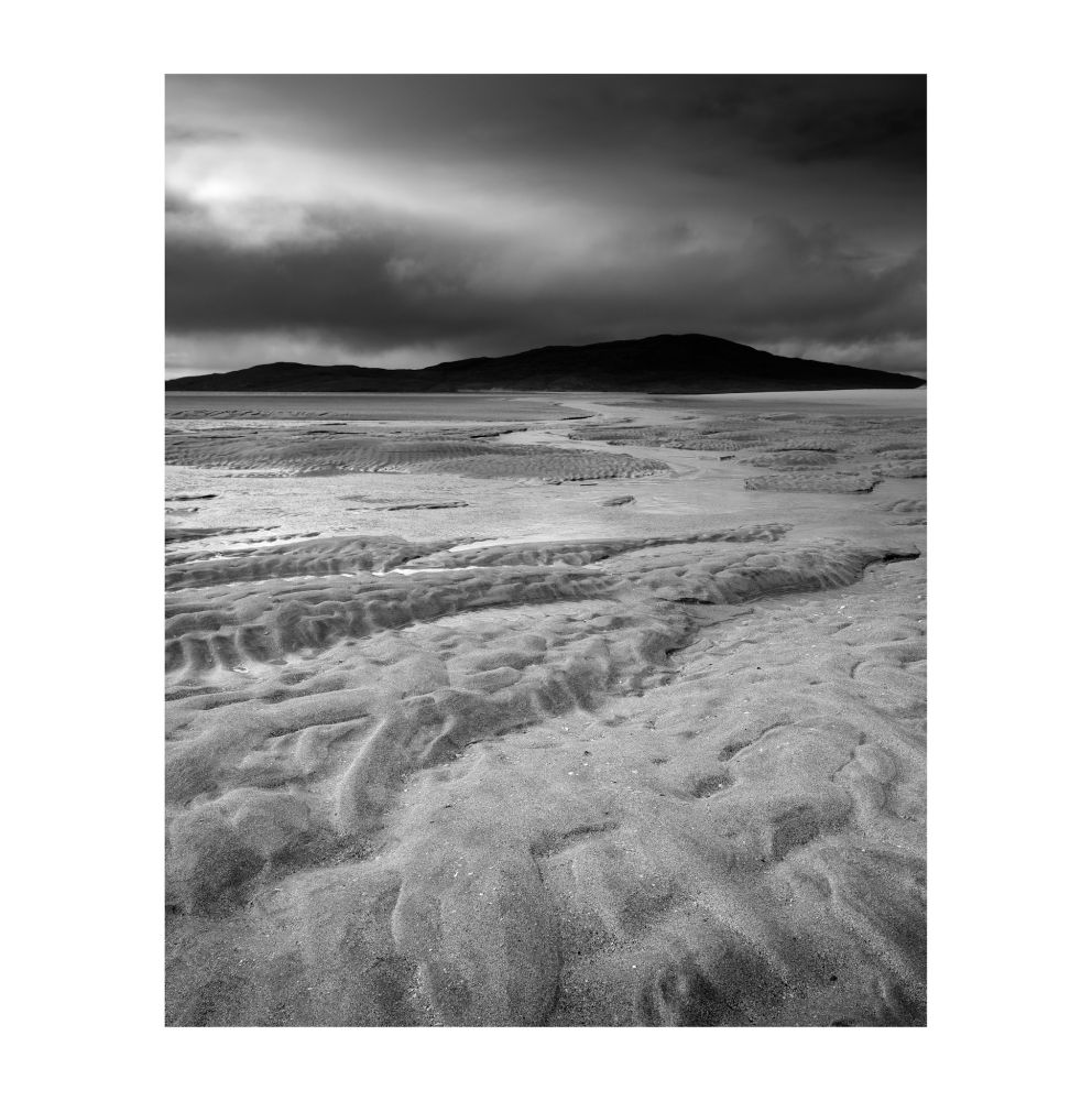 Black and white image of textured sands ripples and snaking tidal waters under the gloom of storm clouds and breaking light, with the silhouette of a distant island on the horizon.