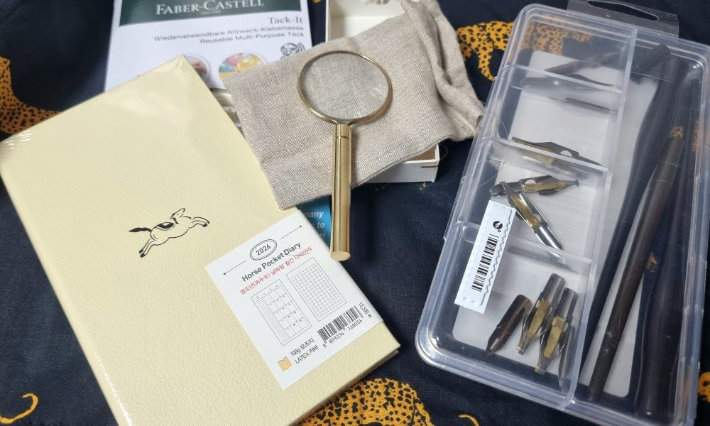 A cream-covered half-sized diary, a magnifying glass with a bronze handle, and dip pen nibs and pen holders in a plastic box on a cheetah-patterned black scarf.