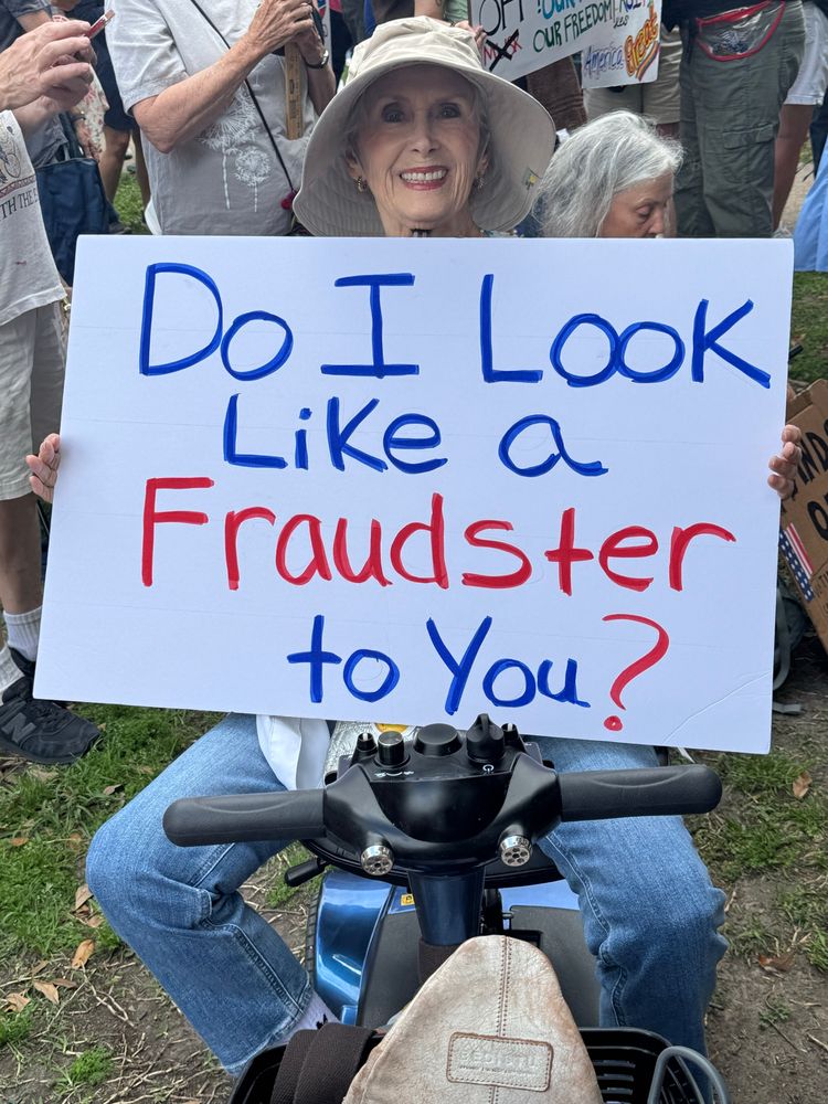 Elderly woman on a mobility scooter holding a protest sign that reads: Do I look like a fraudster to you?