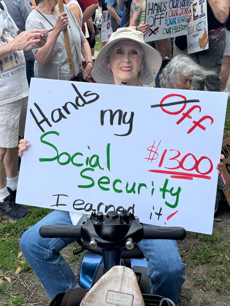 Elderly woman on a mobility scooter holding a protest sign that reads: Hands off my $1300 Social Security. I earned it!