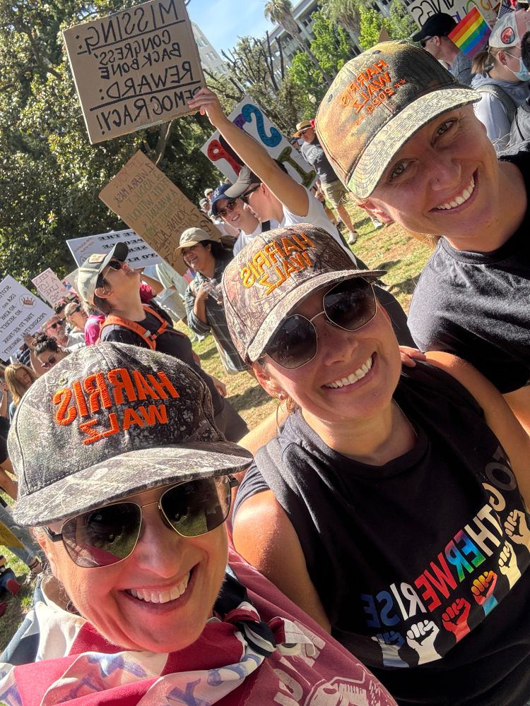 Photo of three women wearing Harris Walz camo hats. 