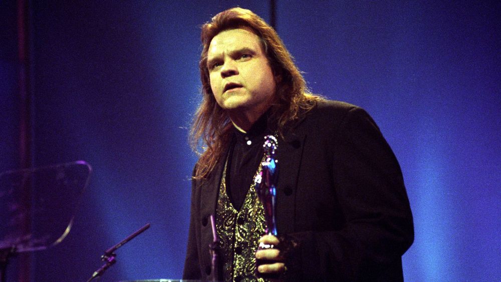 photo of the singer meatloaf with long hair and a suit holding a microphone with a light shining upward behind him