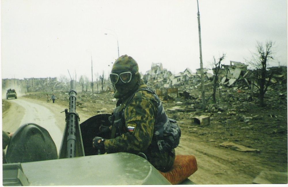 A photograph of a Russian soldier in Grozny c. 2000 looking back at the camera while riding in some sort of military vehicle.
