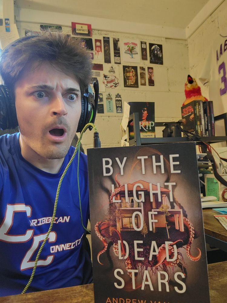 AJ wears headphones that appear to be plugged into a copy of "By The Light of Dead Stars" by Andrew Van Wey
