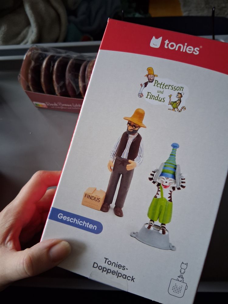 Karton eines 2er Sets Tonies mit Geschichten von Pettersson und Findus:
1. Figur - Pettersson (hat die Geschichte 'Wie Findus zu Pettersson kam')
2. Figur - Findus auf einem Waschtrog, der sich die Ohren zuhält (auf diesem Tonie ist 'Findus und der Hahn im Korb' zu hören)

 Im Hintergrund ist zudem eine Packung Lebkuchen zu sehen