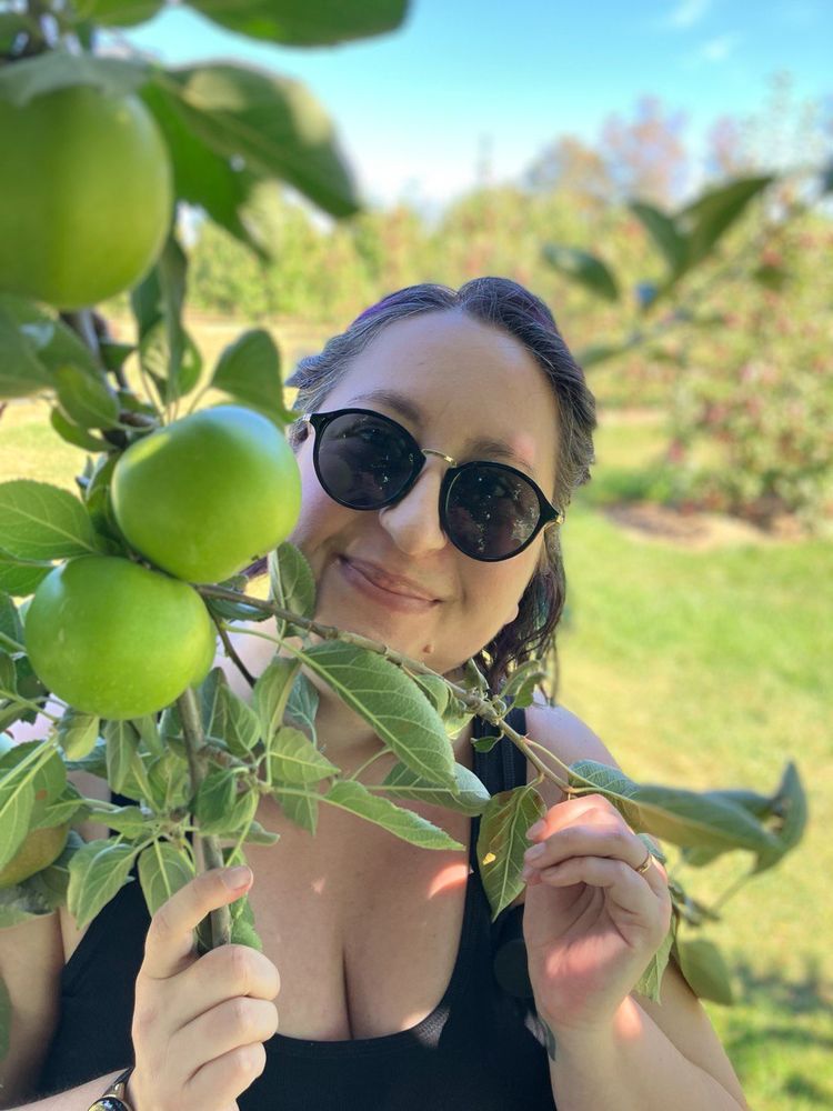 Woman with short dark hair and wear sunglasses is taking a picture in front of a green apple tree. 
