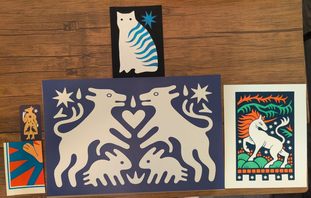 A set of four prints and a business card on a table. They include a cat-like blob with white hair and blue wavy stripes on a black background.There is also a blue pointy star behind the cat. 
There is a business card with a yellow wolf-like creature wearing a dress and white gloves. The wolf is smiling. Inside the wolf's belly is two birds and three blood drops.
There is a cut off portion of a print with red and blue striping and an orange animal's foot. The back side of it says "Thank you so much!"
There is a print of a tapestry with two white dogs and two rabbits on a blue field. They are mirrored down the middle of the print. Also on the tapestry are white flowers, stars, and hearts.
The final print is a whore unicorn with an orange mane and horn. It is standing with one hood raised on a green field. Above the unicorn is a set of green and orange brambles. All of this is set on a dark blue field with white stars dotted throughout. 