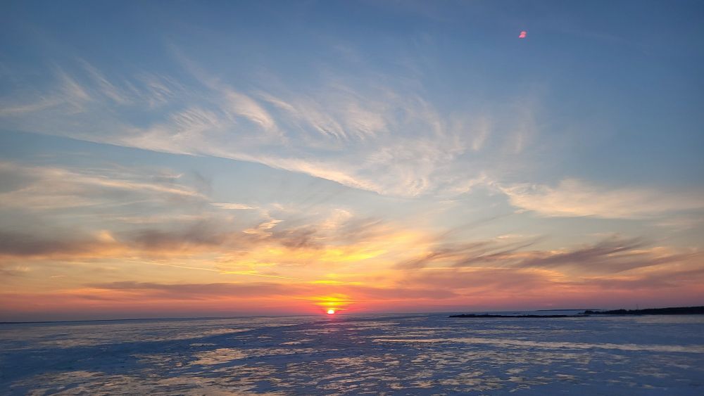Whispy clouds in front of a sky that changes from blue to gold and finishing with red at the horizon creates a beautiful sunset over a frozen Lake Michigan, captured while crossing the Mighty Mackinaw Bridge. (I was not driving)