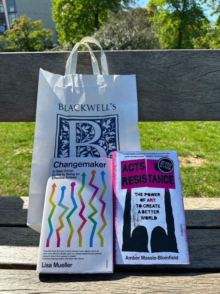 ‘Changemaker: A Data-Driven Guide to Being an Effective Activist’ book and ‘Acts of Resistance: The Power of Art to Create a Better World’ book placed on a bench in a park with a Blackwell’s Oxford bag.