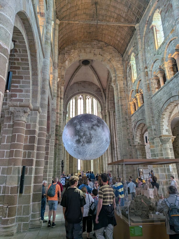 A photo of the interior of the Abbey at Mont-Saint-Michel. There's a crowd, very high ceilings. This is an Abbey, not a cathedral, so it is a very simple interior. Lots of stone, simple glass windows, and a replica of the moon hanging low. The moon is not permanent - it's for a thing they were doing. Moon is still pretty big and impressive. 
