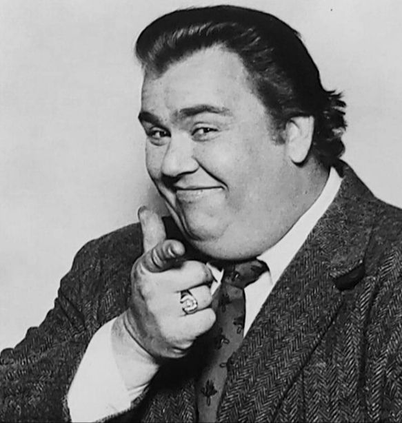 John Candy with the same outfit, but now pointing with his right index finger at the camera and smiling.