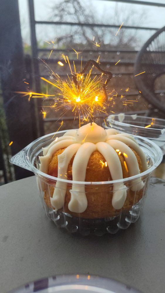 On an outdoor table, a lemon raspberry bundt cake with white frosting with a single star shaped sparkler candle burning and sparking.