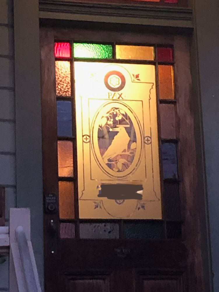 The front door of a house, half of which is glass: multi-colored stained glass framing a large pane engraved with a strikingly similar portrait of a crow delicately holding a berry in its beak. "PAX" is engraved above the crow. 