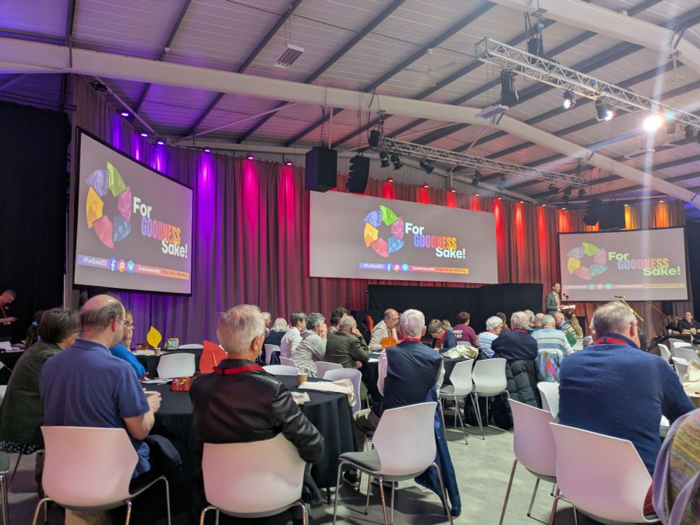 A conference hall with people sitting at round tables and 3 screens with the For Goodness Sake logo. 
