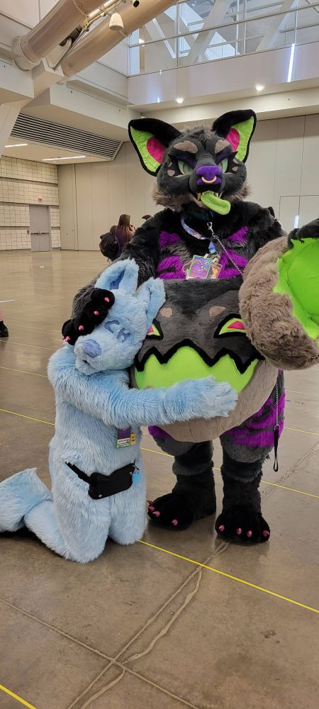 Two fursuiters, YCH and Hyde Hyena, face the camera. YCH is on their knees, wrapping one arm around Hyde's belly as Hyde's right handpaw presses their head against the belly.