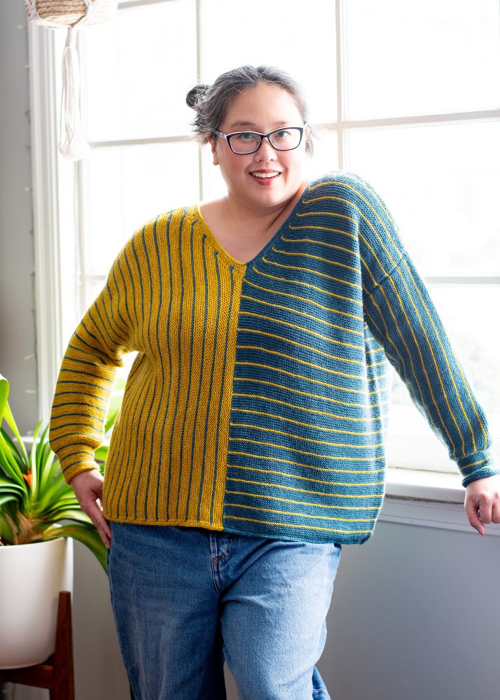 Laura, a Canadian woman of Chinese descent, standing in front of a bright paned window wearing a Perpendicular Pullover in teal and yellow stripes. 