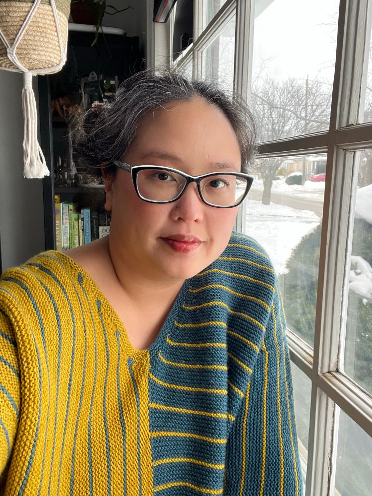 Laura is a Chinese Canadian woman wearing a yellow and teal handknit sweater with horizontal stripes on one half and vertical stripes on the other, with the colours inverted. She’s posing by a window with snow outside. 
