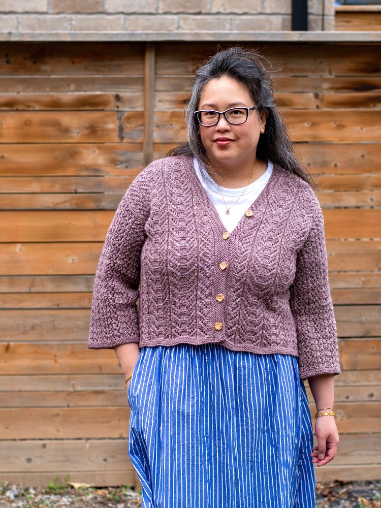 Laura, a Chinese-Canadian woman, poses in front of a wooden fence. She’s wearing a mauve handknit lace cardigan, white tee and blue striped skirt. 