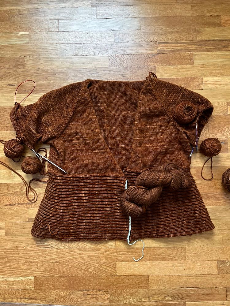 A gingerbread-brown sweater in progress laying on a wooden floor. Both sleeves are on the needles. The lower body of the sweater is worked in perpendicular brioche stitch. 