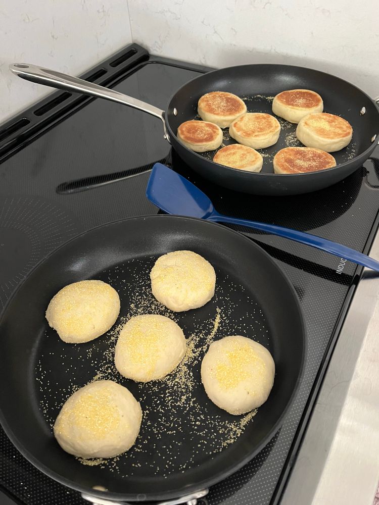 Two dark pans on a stovetop with English muffins in progress. The ones on the back pan are browned and almost done, while the front ones are just getting going. 
