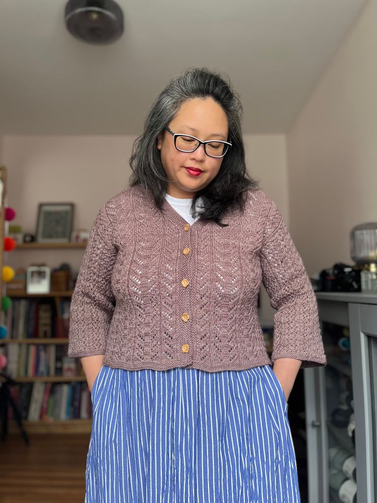 Laura, a Chinese-Canadian woman, wearing a mauve hand knit lace cardigan over a white t-shirt and striped blue skirt. She’s standing in her home studio with her hands in her pockets, looking down. 