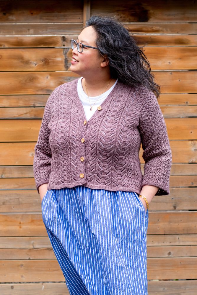 Laura, a Chinese Canadian woman, stands in front of a weathered wood fence. She has her hands in the pockets of a blue and white striped skirt, and is wearing a white tee shirt and mauve handknit lace cardigan with 3/4 sleeves. 
