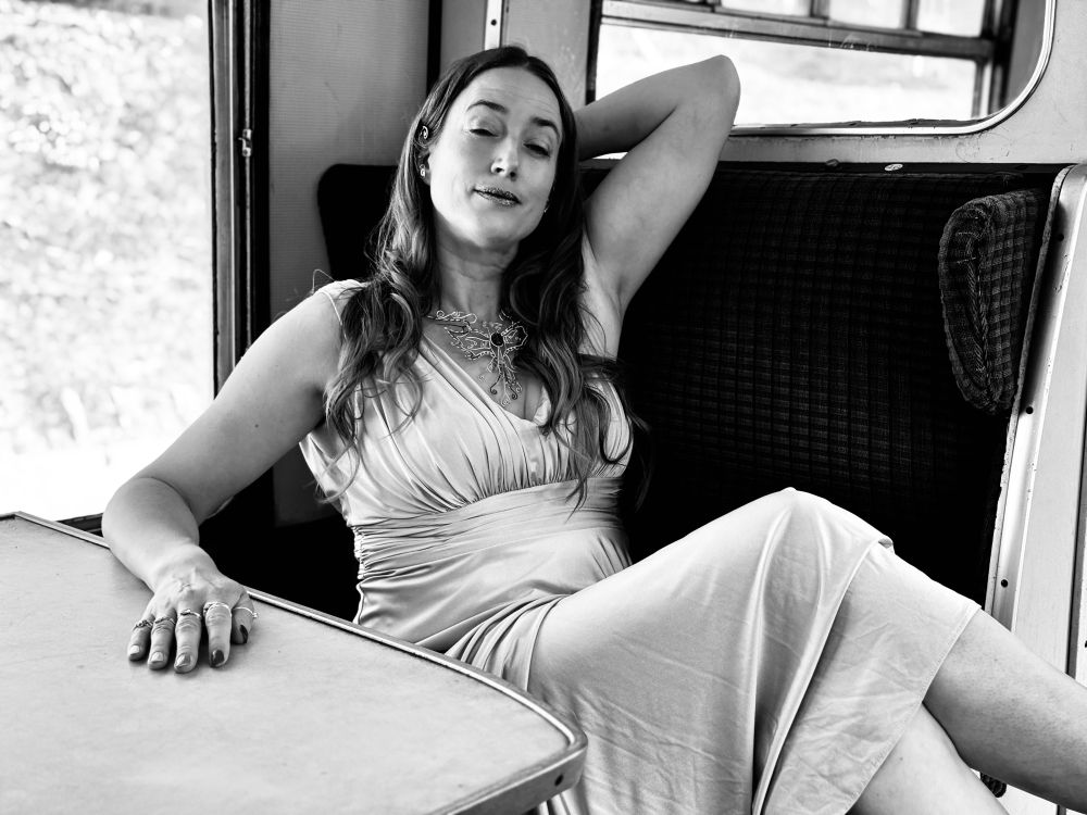 A black and white photo of Ida in a pale satin ballgown, lounging with her eyes closed and legs thrown carelessly over the seat arm inside a steam train carriage.