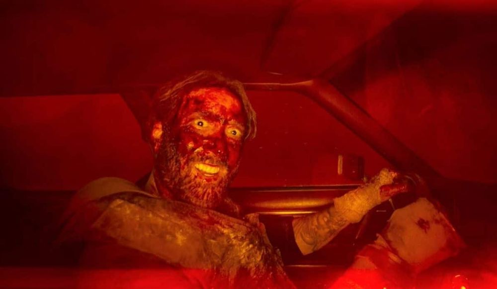 Image: still of Nic Cage from the ending of MANDY, wearing ex stanic motorbiker body armor, covered completely in dry8ng blood and gore, and smiling maniacally into the camera while driving a car. 