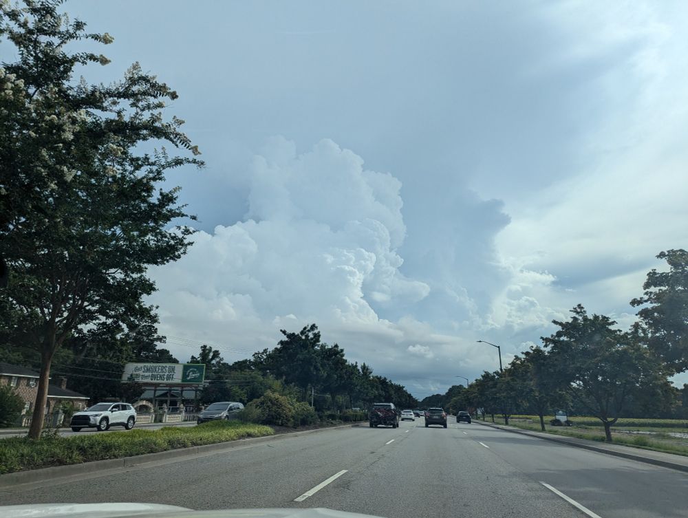 Large cumulous clouds rise in the distance into a ceiling-like layer of flat hazy cloud bases above. Photo is taken from inside a moving car, with a view of the road and green trees taking up the bottom third.